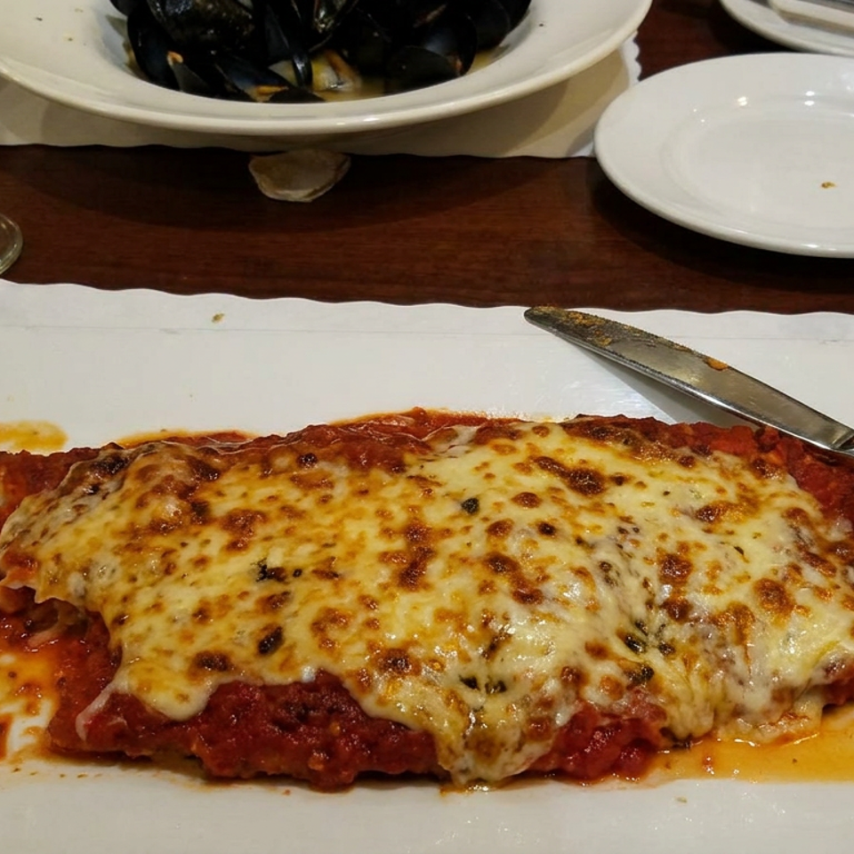 Cheesy chicken parmesan with tomato sauce served on a white plate at a restaurant table, alongside mussels and wine.